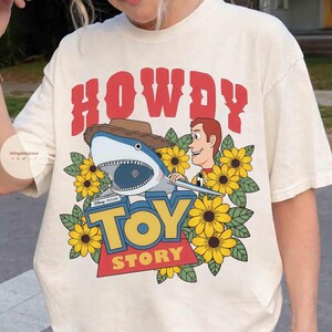 Puede incluir: Camiseta color crema con un dise&ntilde;o gr&aacute;fico. Presenta un tibur&oacute;n con sombrero de vaquero, Woody de Toy Story y las palabras "TOY STORY". El dise&ntilde;o incluye girasoles amarillos y la palabra "HOWDY" en rojo.