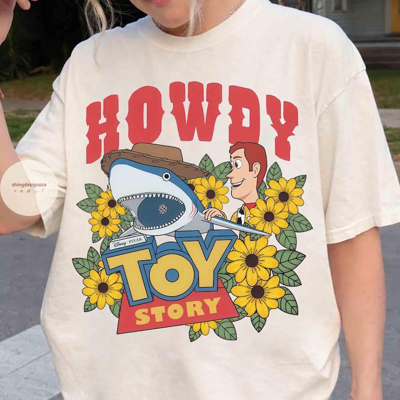Puede incluir: Camiseta color crema con un dise&ntilde;o gr&aacute;fico. Presenta un tibur&oacute;n con sombrero de vaquero, Woody de Toy Story y las palabras "TOY STORY". El dise&ntilde;o incluye girasoles amarillos y la palabra "HOWDY" en rojo.