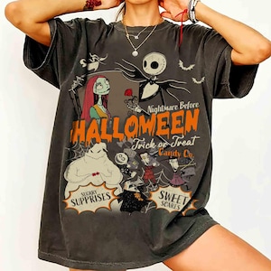 May include: A charcoal gray short-sleeve t-shirt featuring a graphic print of characters from The Nightmare Before Christmas. The design includes the text Nightmare Before Halloween, Trick or Treat, Candy Co., Sugrry Supprises, and Sweet Scares.