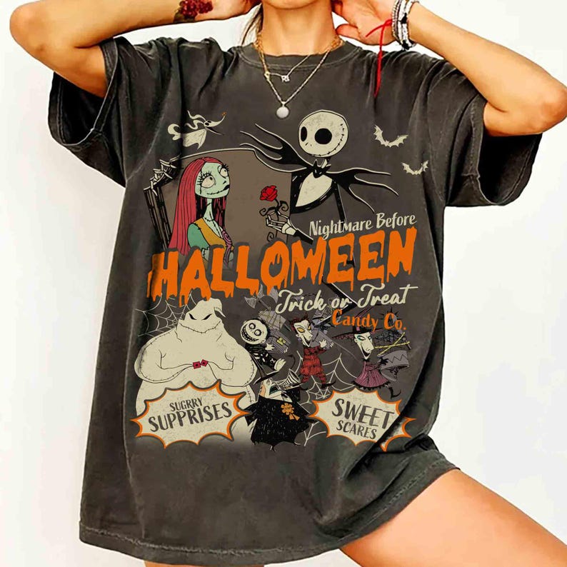 May include: A charcoal gray short-sleeve t-shirt featuring a graphic print of characters from The Nightmare Before Christmas. The design includes the text Nightmare Before Halloween, Trick or Treat, Candy Co., Sugrry Supprises, and Sweet Scares.