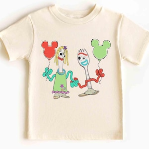 May include: Cream-colored t-shirt featuring a cartoon illustration of two fork-shaped characters holding Mickey Mouse-shaped balloons. One balloon is red, the other is green. The characters are in green and purple outfits.