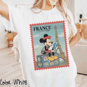 May include: White t-shirt with a graphic of Mickey Mouse in front of the Eiffel Tower, holding a passport. The design is framed like a postage stamp with the word "FRANCE" at the top. The words "Color: White" are at the bottom.
