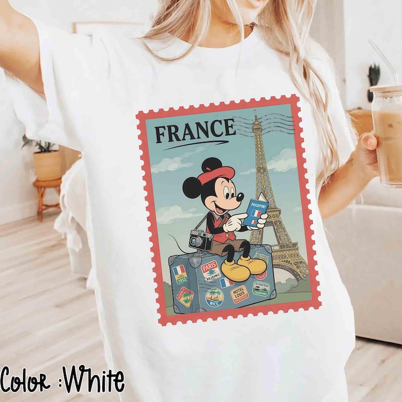 May include: White t-shirt with a graphic of Mickey Mouse in front of the Eiffel Tower, holding a passport. The design is framed like a postage stamp with the word "FRANCE" at the top. The words "Color: White" are at the bottom.