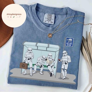 May include: Blue Comfort Colors t-shirt featuring a humorous illustration of Stormtroopers at a bus stop. The design includes an "AT-AT STOP" sign and Stormtroopers with a briefcase, reading, and using a phone. A gold necklace is draped across the shirt.