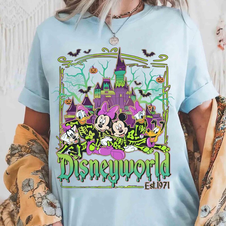 May include: Pale blue t-shirt with a Disney World Halloween design. The graphic shows Disney characters, a castle, bats, and pumpkins. The text "Disneyworld" is in a green, dripping font, with "Est.1971" below.