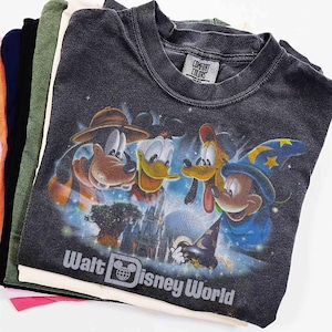 K&ouml;nnte beinhalten: Ein Stapel verschiedenfarbiger T-Shirts. Das oberste Shirt ist dunkelgrau und zeigt eine Grafik der Disney-Figuren Goofy, Donald Duck, Pluto und Mickey Mouse. Der Text "Walt Disney World" ist darunter aufgedruckt.