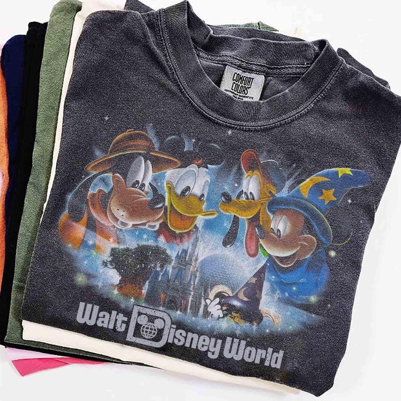 K&ouml;nnte beinhalten: Ein Stapel verschiedenfarbiger T-Shirts. Das oberste Shirt ist dunkelgrau und zeigt eine Grafik der Disney-Figuren Goofy, Donald Duck, Pluto und Mickey Mouse. Der Text "Walt Disney World" ist darunter aufgedruckt.
