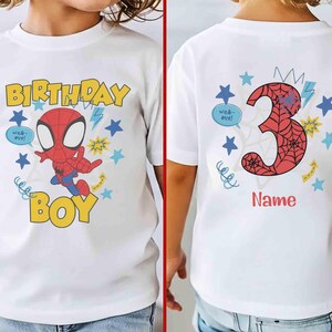 Pu&ograve; includere: Una t-shirt bianca a maniche corte con la grafica di un supereroe dei cartoni animati, il testo "BIRTHDAY BOY" in giallo e un grande numero "3" rosso con motivo a ragnatela con la parola "Name" sotto, circondato da stelle blu.