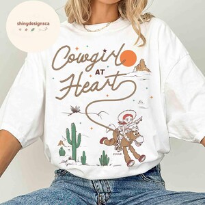 May include: White t-shirt with the text "Cowgirl at Heart" in a rope-style font. The graphic features a cowgirl riding a horse, with a desert landscape and an orange sun in the background. The shirt has a relaxed fit.