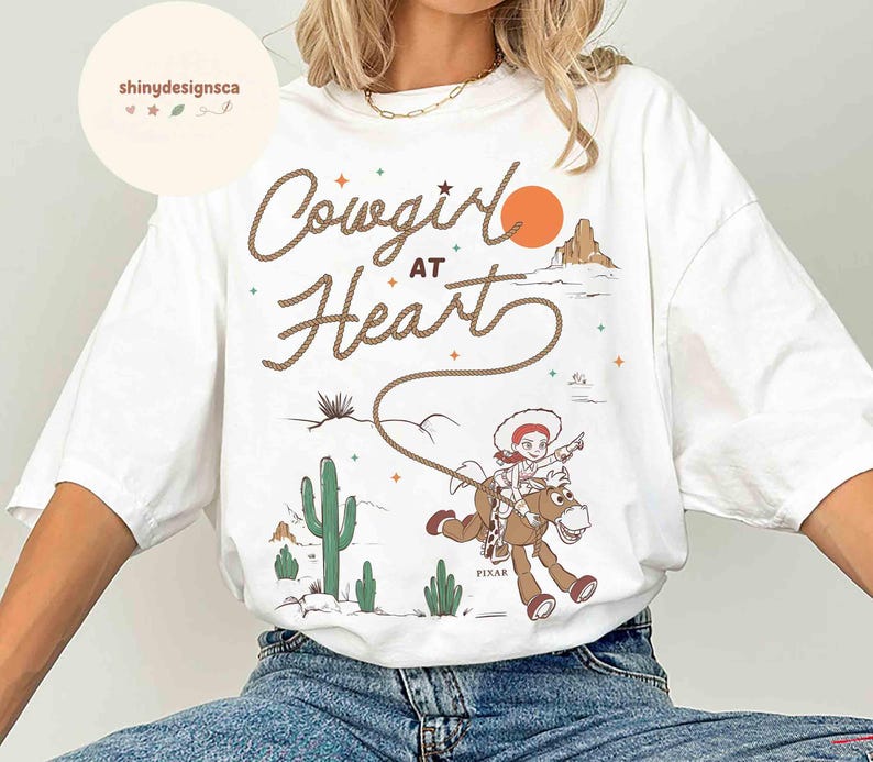 May include: White t-shirt with the text "Cowgirl at Heart" in a rope-style font. The graphic features a cowgirl riding a horse, with a desert landscape and an orange sun in the background. The shirt has a relaxed fit.