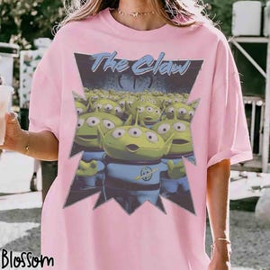 K&ouml;nnte beinhalten: Hellrosa T-Shirt mit einer Grafik von gr&uuml;nen, dre&auml;ugigen Aliens in blauen Uniformen und dem Text "The Claw" in Blau. Das Design ist in einer gezackten schwarzen und grauen Form gehalten.