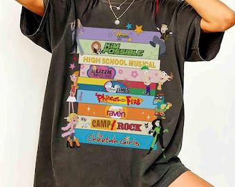 May include: Dark gray t-shirt featuring a colorful graphic of stacked books with cartoon characters and titles like "Kim Possible," "High School Musical," and "Phineas and Ferb." The shirt has a vintage, worn look.