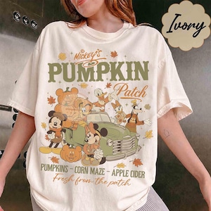 Puede incluir: Una camiseta color marfil con un estampado gráfico de Mickey, Minnie, Donald, Daisy y Goofy con calabazas y un camión vintage. El texto dice Mickey's Pumpkin Patch, Pumpkins, Corn Maze, Apple Cider y Fresh from the patch.