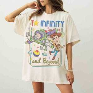 May include: An off-white short-sleeve t-shirt featuring a graphic print of Buzz Lightyear and Woody from Toy Story. The design includes the text "To INFINITY and Beyond" in blue and gold, surrounded by stars, a rocket, and a colorful ball.