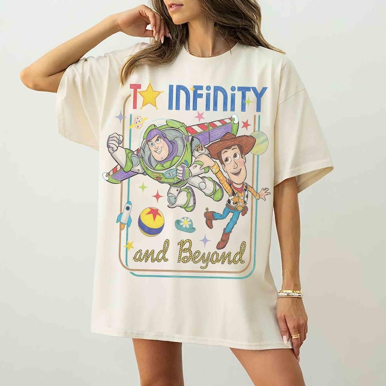 May include: An off-white short-sleeve t-shirt featuring a graphic print of Buzz Lightyear and Woody from Toy Story. The design includes the text "To INFINITY and Beyond" in blue and gold, surrounded by stars, a rocket, and a colorful ball.