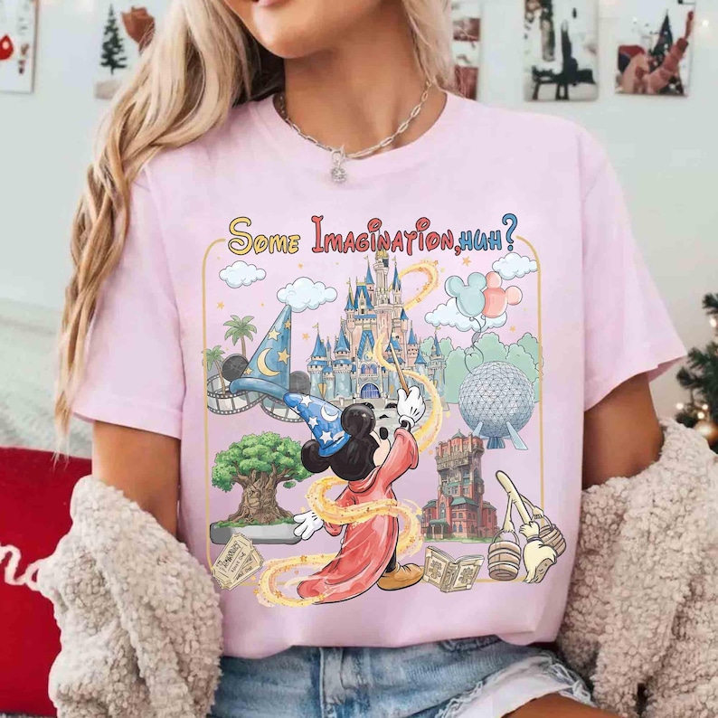 Op de afbeelding: Zachtroze T-shirt met een kleurrijke afbeelding van Mickey Mouse als tovenaar, omringd door themaparkelementen. De tekst "Some Imagination, Huh?" staat erboven. Het shirt heeft korte mouwen.