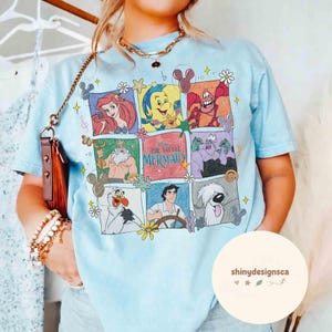 May include: Light blue t-shirt featuring a colorful graphic of characters from Disney's "The Little Mermaid." The design includes Ariel, Sebastian, Flounder, King Triton, Ursula, Olaf, Prince Eric, and Max. The shirt has a crew neck and short sleeves.