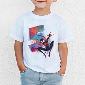 May include: White t-shirt with a colourful graphic of the number 5 and a Spider-Man character in a dynamic pose. The design incorporates red, blue, and black hues, creating a vibrant and playful look. The shirt is likely for a child.