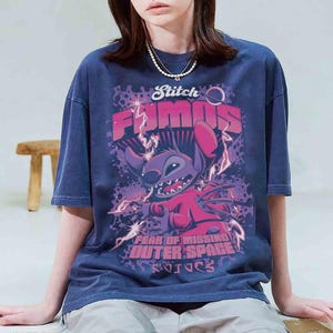 May include: Navy blue t-shirt featuring a graphic of Stitch from Lilo & Stitch. The design includes the words "Stitch Famous" and "Fear of Missing Outer Space" in pink and purple. The shirt has a vintage wash.