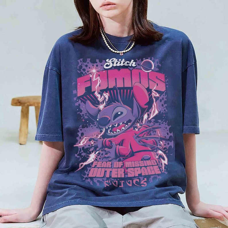 May include: Navy blue t-shirt featuring a graphic of Stitch from Lilo & Stitch. The design includes the words "Stitch Famous" and "Fear of Missing Outer Space" in pink and purple. The shirt has a vintage wash.