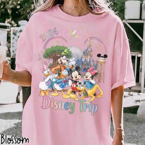 May include: A light pink t-shirt featuring a colorful graphic of cartoon characters including Mickey Mouse, Minnie Mouse, Donald Duck, Daisy Duck, Goofy, and Pluto. The design includes the text 2026 and Disney Trip, a castle, and a water tower.