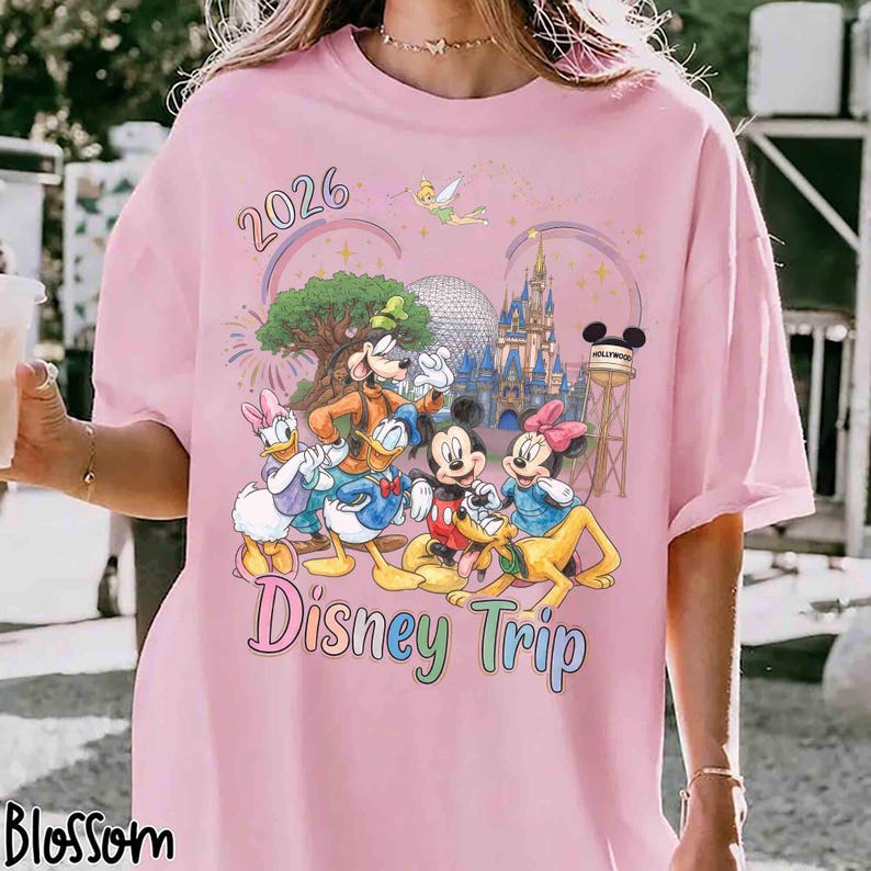 May include: A light pink t-shirt featuring a colorful graphic of cartoon characters including Mickey Mouse, Minnie Mouse, Donald Duck, Daisy Duck, Goofy, and Pluto. The design includes the text 2026 and Disney Trip, a castle, and a water tower.