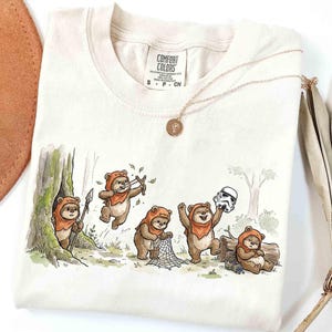 May include: Cream-colored t-shirt featuring a whimsical illustration of Ewoks in a forest setting. The design includes Ewoks with a slingshot, net, and Stormtrooper helmet. A gold necklace with a pendant rests on the shirt.