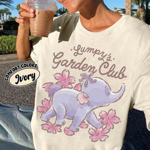 Puede incluir: Camiseta color marfil con el texto "Lumpy's Garden Club" y una ilustración de elefante de dibujos animados. El elefante es de color púrpura claro con detalles en rosa y rodeado de flores rosas. La camiseta también tiene las palabras "Comfort Colors" e "Ivory".