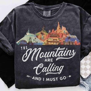 May include: A charcoal gray short-sleeve t-shirt featuring a colorful graphic of various mountain-themed amusement park attractions. The text reads "THE Mountains ARE Calling AND I MUST GO" in white script and block lettering with arrow accents.