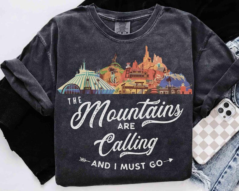May include: A charcoal gray short-sleeve t-shirt featuring a colorful graphic of various mountain-themed amusement park attractions. The text reads "THE Mountains ARE Calling AND I MUST GO" in white script and block lettering with arrow accents.