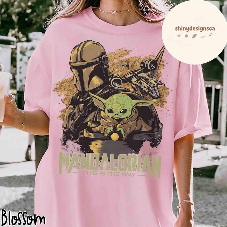 Puede incluir: Camiseta rosa claro con un dise&ntilde;o de The Mandalorian y Grogu, en tonos dorados y marrones. El texto "MANDALORIAN" y "THIS IS THE WAY" est&aacute; impreso en verde. La camiseta tiene un corte holgado.