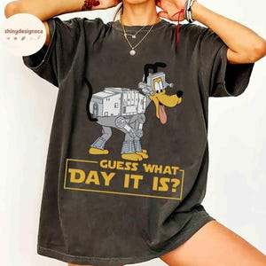 Może przedstawiać: Ciemnoszara koszulka z kreskówkowym psem w roboczym projekcie AT-AT. Pies ma długi język i nosi słuchawki. Tekst "GUESS WHAT DAY IT IS?" znajduje się w żółtym prostokącie.