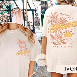 May include: Ivory-colored t-shirt featuring a tropical design with the words "Ohana Beach" and "Enjoy Life". The design includes palm trees, a sunset, and a cartoon character. The shirt is a relaxed fit.