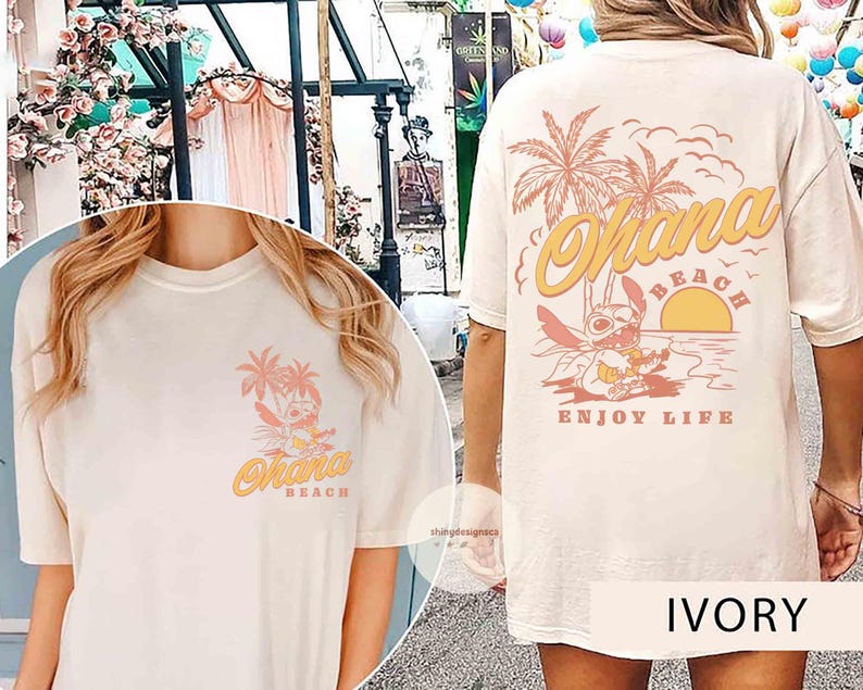 May include: Ivory-colored t-shirt featuring a tropical design with the words "Ohana Beach" and "Enjoy Life". The design includes palm trees, a sunset, and a cartoon character. The shirt is a relaxed fit.