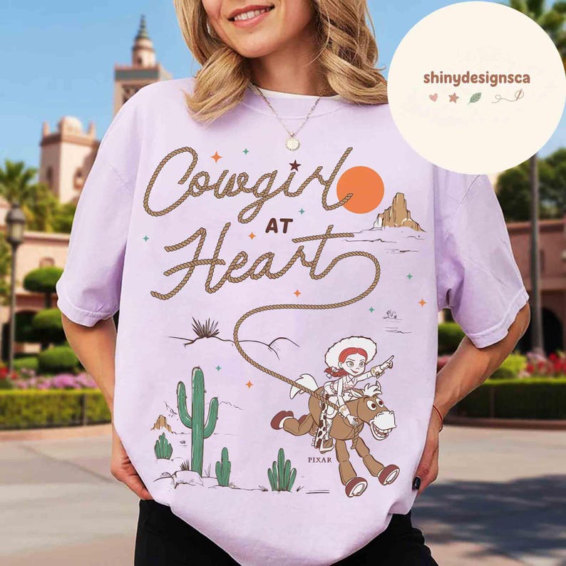 May include: A light purple t-shirt with the words "Cowgirl at Heart" in a rope-like font, featuring a cartoon cowgirl riding a horse. The design includes a desert landscape with cacti and a setting sun. The word "PIXAR" is at the bottom.