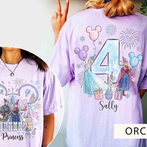 May include: A light purple t-shirt featuring a graphic print of two animated characters, a snowman, and a large number 4. The design includes balloons, fireworks, and gift boxes. The name Sally is printed below the graphic in a cursive font.