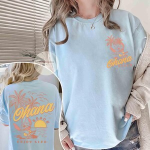 May include: Light blue t-shirt with a tropical design. The front features a palm tree and the word "Ohana" in orange. The back of the shirt has a larger design with a cartoon character, palm trees, and the words "Ohana Beach Enjoy Life".