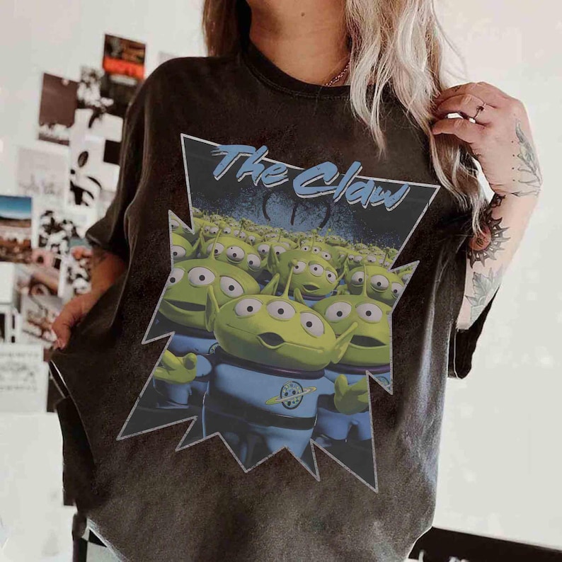 K&ouml;nnte beinhalten: Dunkelgraues T-Shirt mit einer Grafik von gr&uuml;nen Aliens aus dem Film Toy Story. Die Aliens tragen blaue Uniformen und stehen vor einem blau-schwarzen Hintergrund. Der Text "The Claw" steht &uuml;ber den Aliens.