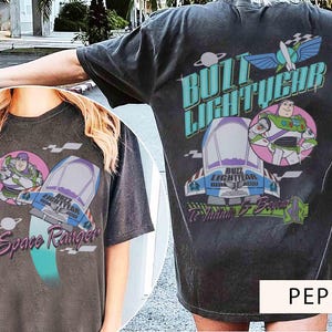 May include: A charcoal gray t-shirt featuring a graphic of Buzz Lightyear, a spaceship, and the text Buzz Lightyear, Space Ranger, and To Infinity & Beyond. The design includes a planet, wings, and a checkered flag pattern in teal, purple, and white.