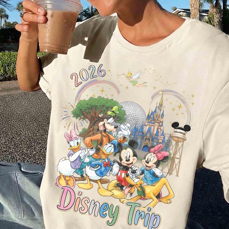 May include: A cream-colored t-shirt featuring a colorful graphic of Disney characters including Mickey Mouse, Minnie Mouse, Donald Duck, Daisy Duck, and Goofy. The design includes the text 2026 and Disney Trip, a castle, a geodesic dome, and Tinker Bell.