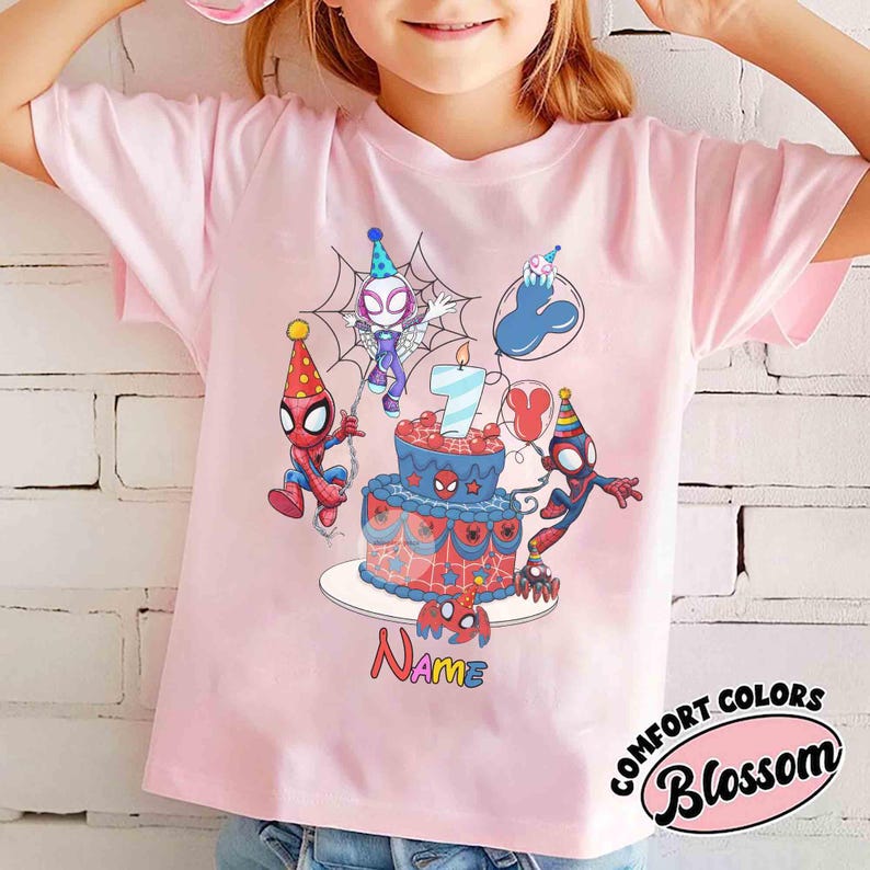 May include: A light pink t-shirt featuring a colorful graphic of Spider-Man characters celebrating a birthday. The design includes a cake with a lit candle, balloons, and the number 7. The word "Name" is written below the cake.