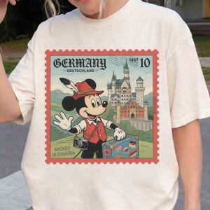 May include: A cream-coloured t-shirt with a postage stamp design. The stamp features Mickey Mouse in Bavarian dress, the word "Germany", and a castle. The text "Mickey in Bavaria" is also visible.