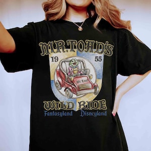 K&ouml;nnte beinhalten: Schwarzes T-Shirt mit einem Aufdruck von Mr. Toad's Wild Ride aus Disneyland. Das Design zeigt ein rotes Auto mit Mr. Toad, den Text "Mr. Toad's Wild Ride" und die Jahreszahlen 19 und 55. Das Shirt ist ein klassischer Stil.