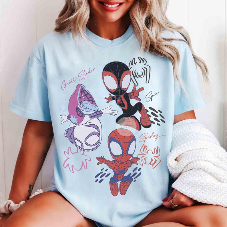 Op de afbeelding: Lichtblauw T-shirt met cartoon illustraties van Spider-Man personages, waaronder Ghost Spider, Miles Morales en Spider-Man. Het shirt heeft de woorden "Ghost Spider", "Spin" en "Spidey" in een handschrift.