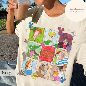 May include: Ivory-colored t-shirt featuring a colorful comic-style illustration of Peter Pan characters. The design includes Peter Pan, Tinkerbell, Captain Hook, and the crocodile. The text "Peter Pan" is visible in the center of the design.