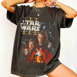 May include: Dark gray t-shirt featuring a Star Wars movie poster design. The graphic includes the text "Star Wars Revenge of the Sith" and images of characters like Darth Vader, Anakin Skywalker, and Obi-Wan Kenobi. The shirt has a vintage, worn look.