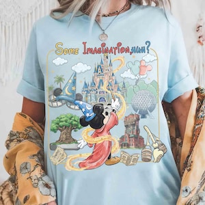 Op de afbeelding: Lichtblauw T-shirt met een afbeelding van Mickey Mouse als tovenaar, die een toverstaf gebruikt en themapark-herkenningspunten. De tekst "Some Imagination, Huh?" staat boven de afbeelding. De mouwen zijn opgerold.