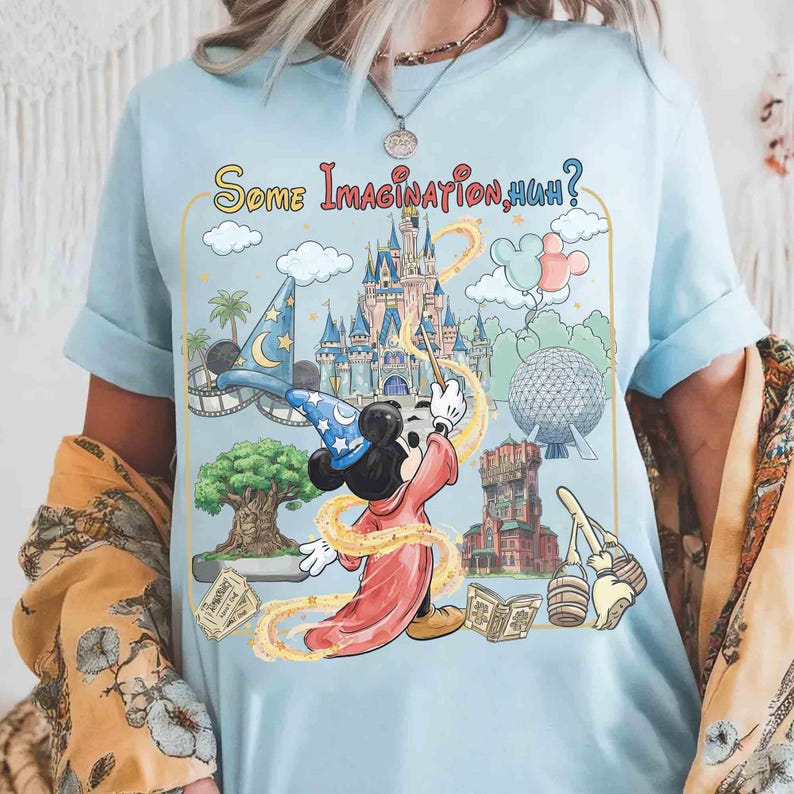 Op de afbeelding: Lichtblauw T-shirt met een afbeelding van Mickey Mouse als tovenaar, die een toverstaf gebruikt en themapark-herkenningspunten. De tekst "Some Imagination, Huh?" staat boven de afbeelding. De mouwen zijn opgerold.