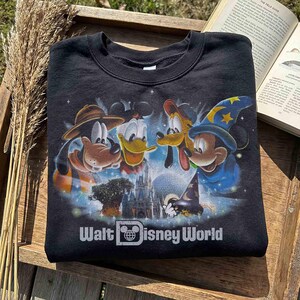 K&ouml;nnte beinhalten: Schwarzes Sweatshirt mit einem farbenfrohen Grafikdesign von Mickey Mouse, Donald Duck, Goofy und Pluto, mit einem Schloss und dem Text "Walt Disney World". Das Design ist detailliert vor einem Sternenhintergrund.