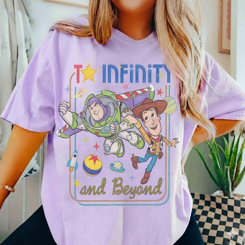 May include: A light purple t-shirt featuring a graphic print of Buzz Lightyear and Woody from Toy Story. The design includes the text "INFINITY and Beyond" with colorful stars, a rocket, and a ball, all enclosed within a rectangular border.